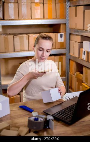 Female small business owner using mobile app on smartphone checking parcel box. Warehouse worker, seller holding phone scanning retail dropshipping bo Stock Photo