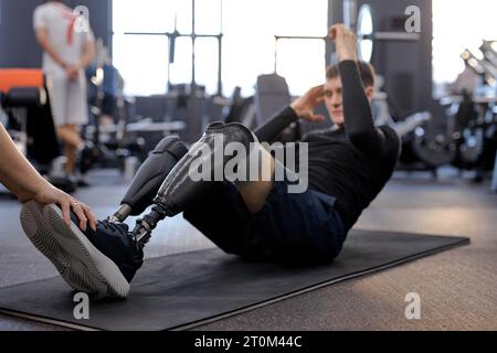 motivated strong man with legs prosthesis doing abdominal crunches, disabled man working out at gym. full length side view shot doing ABS, focus on ar Stock Photo