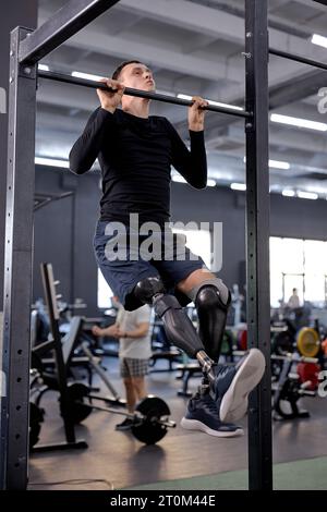 active ambitious man with prosthetic leg wearing sports clothing doing pull-ups on bar in modern gym, full length side view shot, free time, spare tim Stock Photo