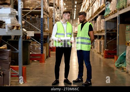 Men warehouse workers talking. holding document paper. two young workers in green uniform discuss together. Concept of good management system to suppo Stock Photo