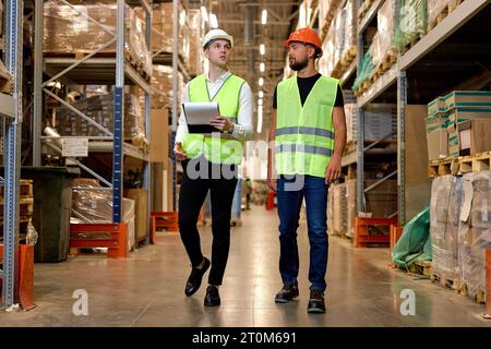 Caucasian engineers in helmets and uniform, team checking goods and supplies on shelves in warehouse. logistic and business export. good management sy Stock Photo