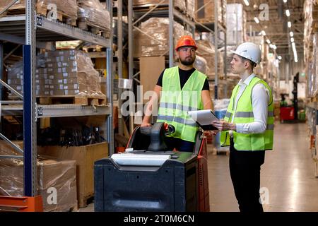 Young caucasian men workers doing stocktaking of products on shelves in warehouse together, using clipboard and forklift, having talk discussing, cont Stock Photo