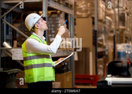 concentrated caucasian man worker in hard hat and working wear outfit doing stocktaking of products on shelves in warehouse using clipboard, taking no Stock Photo