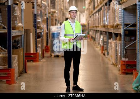 nice caucasian man worker in hard hat and working wear outfit doing stocktaking of products on shelves in warehouse using clipboard, taking notes. Phy Stock Photo