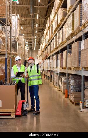 two men warehouse workers with hand pallet jack lift stand having talk, temwork. hand pallet truck and male mover. Workers in uniform with cargo cart. Stock Photo