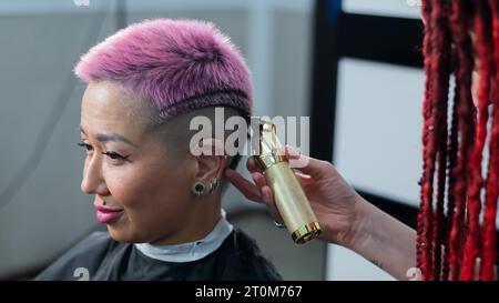 The hairdresser shaves the temple of a female client. Asian woman with short pink hair in barbershop. Stock Photo