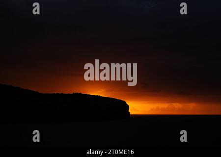 Atmospheric warm glow of sea sunrise through blackness of sky, sea, and cliffs. High contrast low-key minimalist image taken on the south coast of Mal Stock Photo