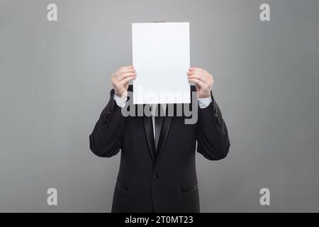 Business person wearing black suit hiding his face behind blank empty white paper on gray studio wall background Stock Photo