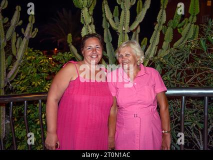 Mother and daughter in bright red dresses are posing near metal handrail on night 5th Centennial Avenue, Puerto de Santiago, Tenerife. Stock Photo