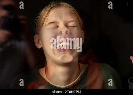 Portrait of a teenage girl through her face pressed against the glass on a dark background. Funny antics Stock Photo