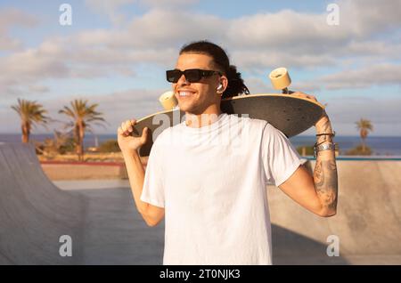 Happy young tattooed man holding skateboard on shoulders outdoors Stock Photo