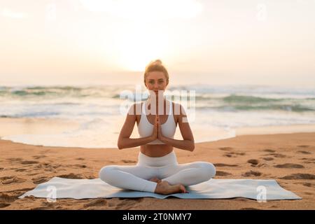 Sunset meditation. Lady meditating outdoors on sea beach, sitting on fitness mat and keeping hands in namaste gesture Stock Photo