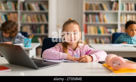 Smiling european schoolgirl at desk with laptop, writing in copybook Stock Photo