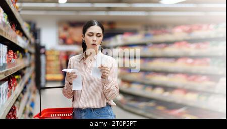 Web banner of shopping and high price. Portrait of shocked Caucasian woman holds paper receipts of purchase. In background, rows of shelves with produ Stock Photo