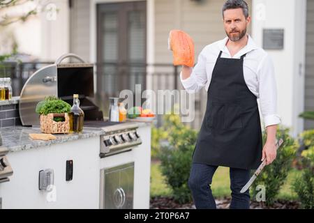 Handsome man in shirt and in cook apron preparing fish on barbecue. Male cook cooking salmon fillet on grill Stock Photo