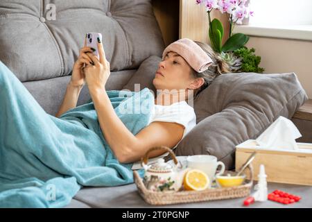 High temperature. A sick woman with a small towel on forehead lies under a blanket and holds a smartphone in her hands. In front of her there is a tra Stock Photo