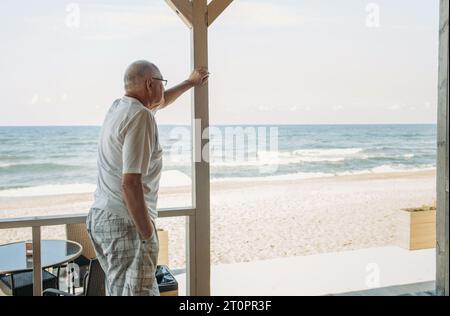 Portrait of a pensive elderly man standing on the terrace of a cafe and looking at the sea. Stock Photo