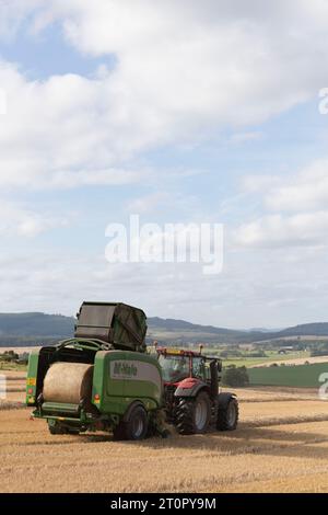 Rear View of a McHale Baler Towed by a Red Valtra Tractor Producing a Straw Bale, with Views Over the Surrounding Aberdeenshire Countryside Stock Photo