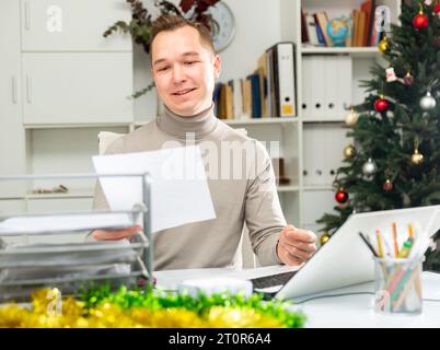 Positive man bookkeeper doing paperwork in office during Christmastime Stock Photo