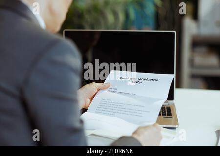 New job. Seen from behind modern middle aged woman worker in modern green office in grey business suit with laptop and employee termination letter. Stock Photo