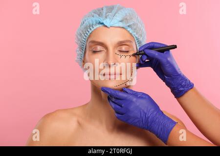 Woman preparing for cosmetic surgery, pink background. Doctor drawing markings on her face, closeup Stock Photo