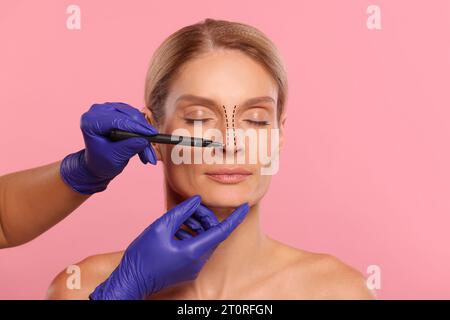 Woman preparing for cosmetic surgery, pink background. Doctor drawing markings on her face, closeup Stock Photo