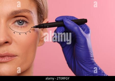 Woman preparing for cosmetic surgery, pink background. Doctor drawing markings on her face, closeup Stock Photo