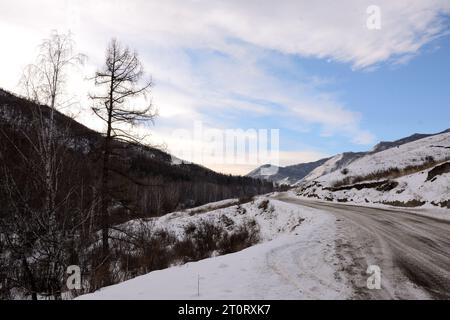 A gravel road covered with snow going through a sparse forest along the slope of a high mountain on a sunny winter day. Altai, Siberia, Russia. Stock Photo