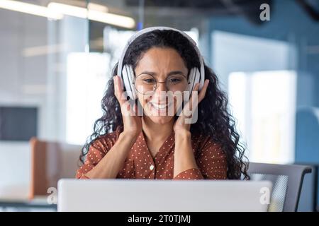 Young beautiful woman in headphones working with laptop inside office at workplace, joyful and satisfied business woman watching online webinar, educational video course, learning remotely. Stock Photo