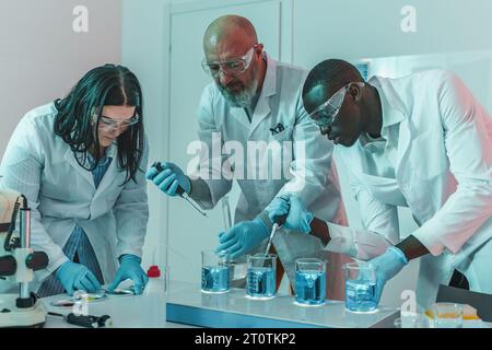 Two young scientists, one caucasian and one African, along with a senior Caucasian colleague, use beakers on a hot plate with a magnetic stirrer in a Stock Photo