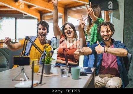 A group of four content creators jubilantly celebrate and gesture towards a smartphone mounted on a tripod, with a microphone in front of them, as the Stock Photo