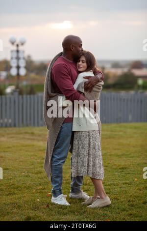 Stunning diverse couple standing in park, embraced in love, wrapped in warm blanket. Affectionate moment between African American man and Caucasian wo Stock Photo