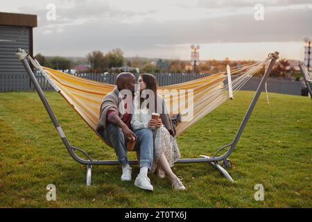 Loving couple kissing sitting in hammock outdoors, wrapped in warm blanket and holding cups with beverage in hands. Black man and Caucasian woman in l Stock Photo