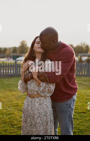 Portrait of interracial couple in love, gently hugging and kissing with eyes closed in nature park. Man embrace girlfriend from behind, standing toget Stock Photo