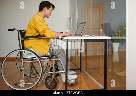 Young man in wheelchair and working on laptop in office of IT company Stock Photo