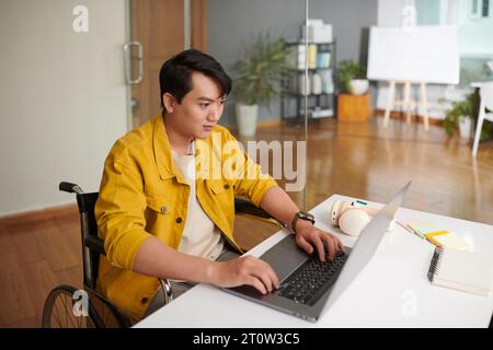 Vietnamese software developer in wheelchair working on laptop in office Stock Photo