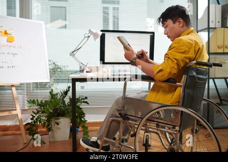 Serious young entrepreneur in wheelchair reading document or article on tablet computer Stock Photo