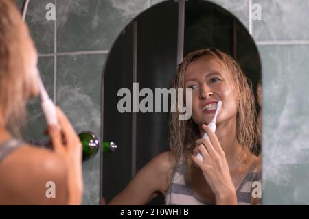 Young blonde woman is brushing her teeth with a white electric toothbrush in the bathroom looking in the mirror. Hygiene, the concept of body care. Stock Photo