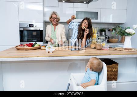 Grandmother mother and baby granddaughter cooking together in kitchen in apartment while mom making a funny faces for child and granny cutting vegetab Stock Photo