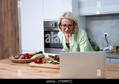 Senior adult woman cooking healthy food for dinner in her domestic kitchen using laptop computer to read online recipe. Grandmother preparing vegetabl Stock Photo