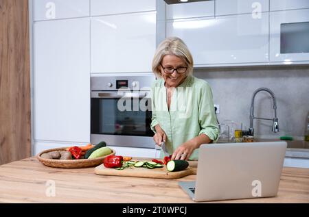 Senior adult woman cooking healthy food for dinner in her domestic kitchen using laptop computer to read online recipe. Grandmother preparing vegetabl Stock Photo
