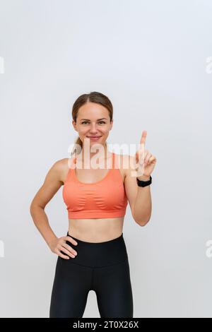 Woman fitness trainer wearing sportswear posing in front of white backdrop, smiling and pointing with finger up. Stock Photo