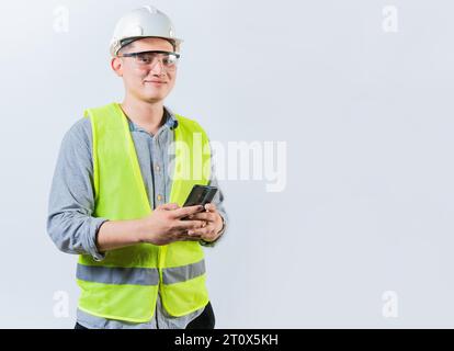 Smiling engineer holding phone looking at camera. Latin male engineer using phone isolated. Young engineer in vest and helmet using cell phone Stock Photo