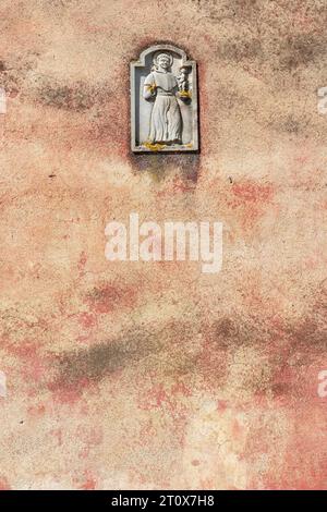 Somewhat dirty and weathered plastered façade or wall of a building in Italy with on top a religious monk and child sculptural plaque Stock Photo