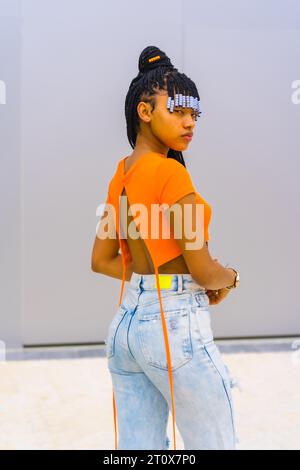 A young trap dancer with braids. Black ethnic girl with orange t-shirt and cowboy pants. Pose with his back Stock Photo