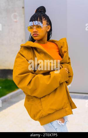 A young trap dancer with braids. Black ethnic girl with t-shirt, orange sunglasses and a jacket, urban post looking at the left Stock Photo