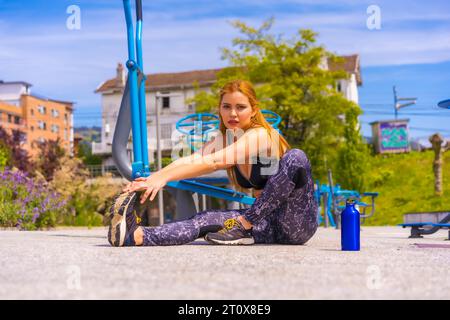 Red-haired woman with purple leggings and black overalls exercising on the machines, sport in the city Stock Photo
