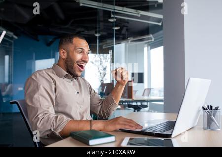Successful businessman celebrating successful completion of project, man holding hands raised received online notification of triumph and good achievement results on laptop, working inside office. Stock Photo