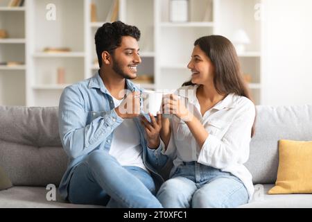 Happy indian couple talking and drinking coffee, sitting on sofa Stock Photo
