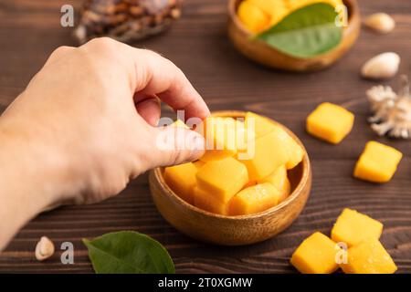 Dried and candied mango cubes in wooden bowls on brown wooden textured background with hand. Side view, close up, selective focus, vegan, vegetarian f Stock Photo